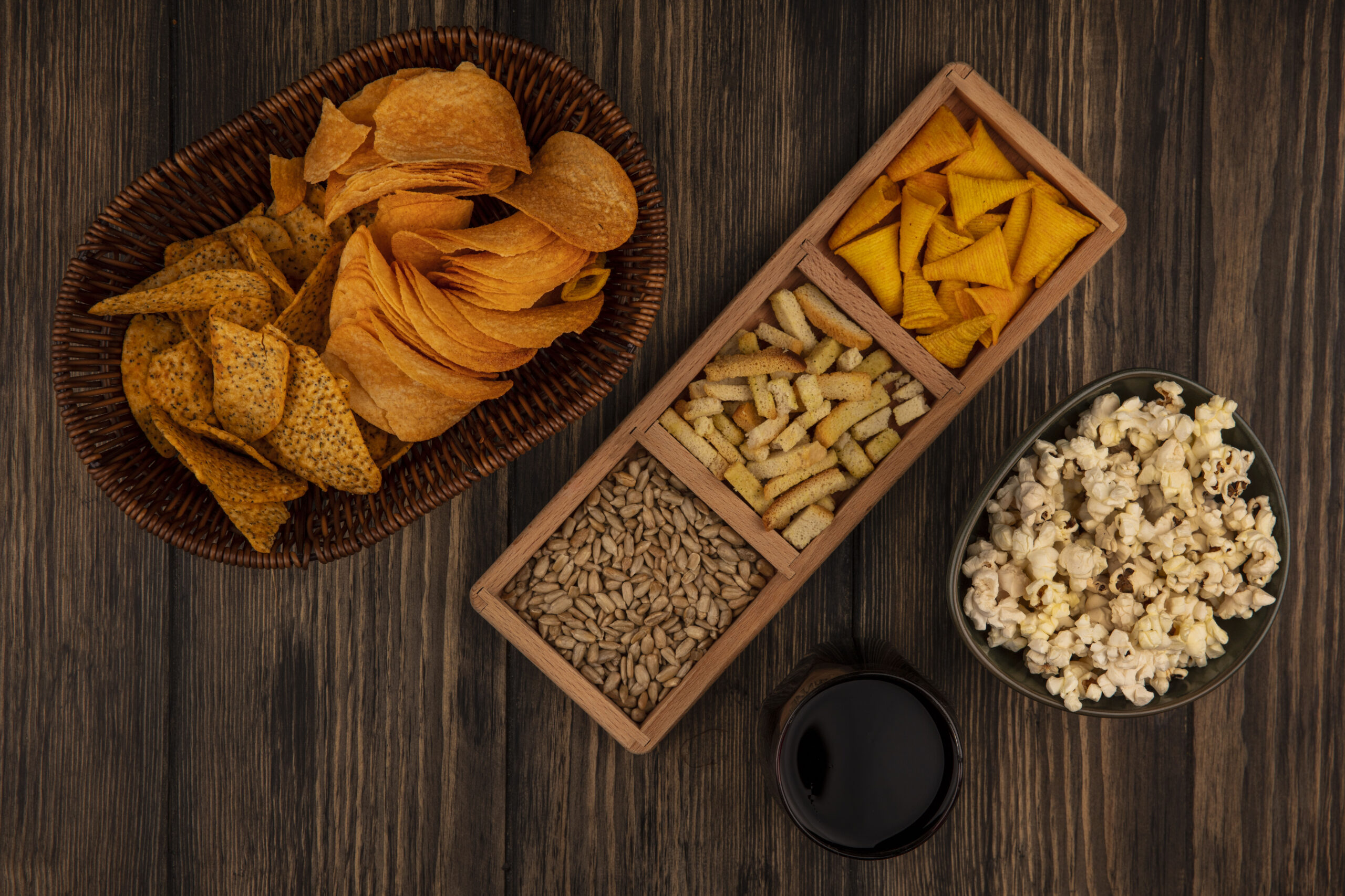 top view of cone shape bugles chips on a wooden divided plate with shelled sunflower seeds with spicy chips on a bucket with a glass of cola on a wooden background